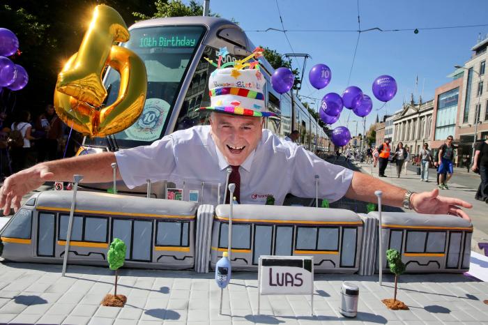 Image from the Luas 10 year anniversary. Image of man in party hat with a Luas style cake in front of the Luas on St Stephens Green.